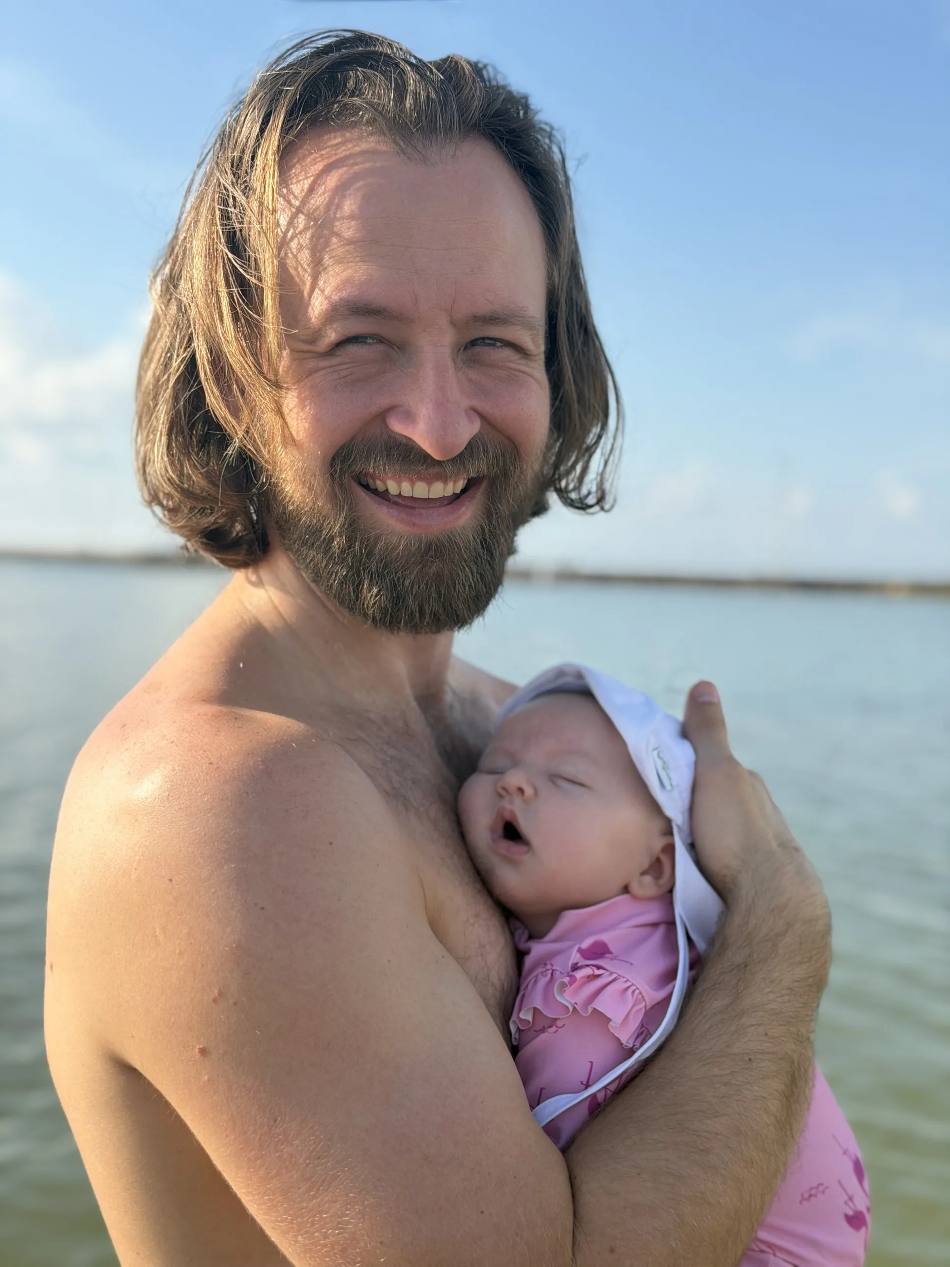 Andrew smiling and holding his infant daughter outdoors by the water